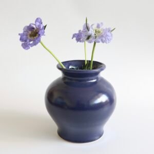 Vintage donkerblauwe aardewerk vaas met paarse bloemen en groene stengels.