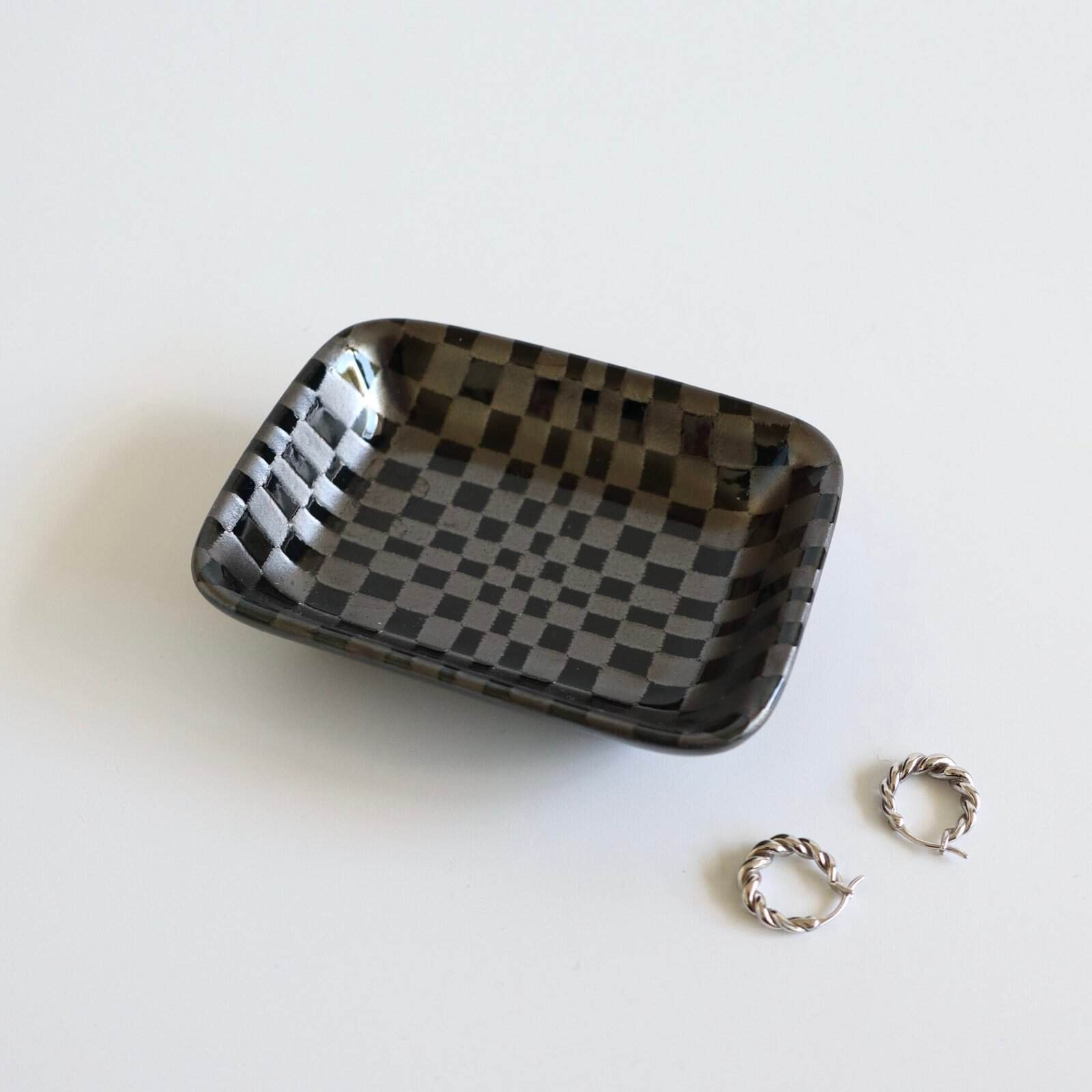 Mini catch-all schaaltje met zwart-grijs geruit patroon, naast twee zilveren ringen op wit oppervlak.