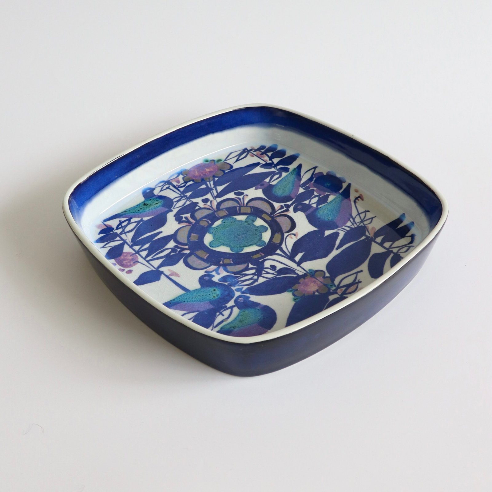Vintage Royal Copenhagen fajance schaal met afgeronde vierkante vorm, blauw-witte glazuur en botanisch decor in donkerblauw met gekleurde accenten.
