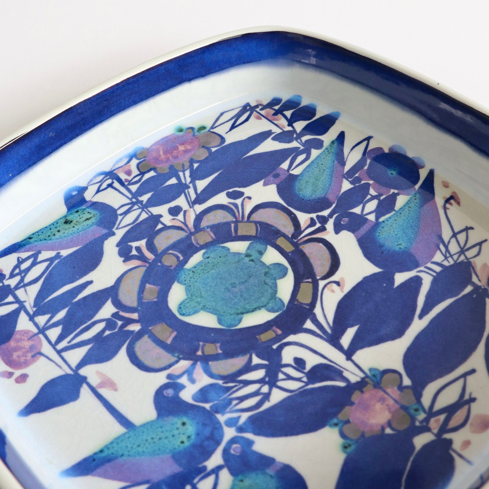 Detailweergave van vogel en bloemmotief op Royal Copenhagen schaal met blauw en paarse accenten.